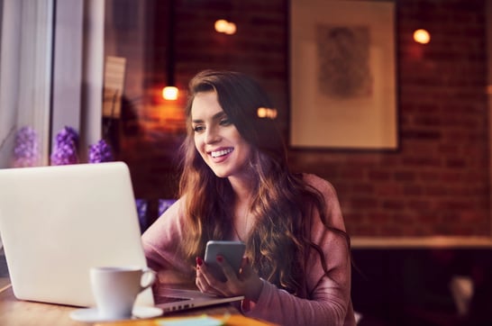 Girl on laptop looking at laptop, holding phone in cafe